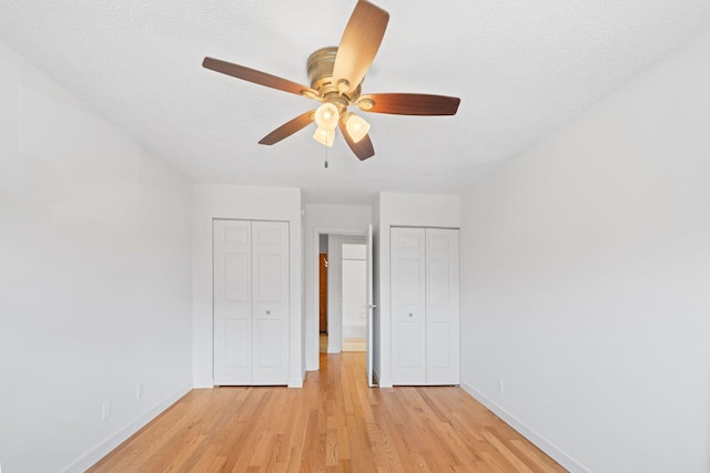 unfurnished bedroom featuring a textured ceiling, ceiling fan, baseboards, light wood-type flooring, and two closets