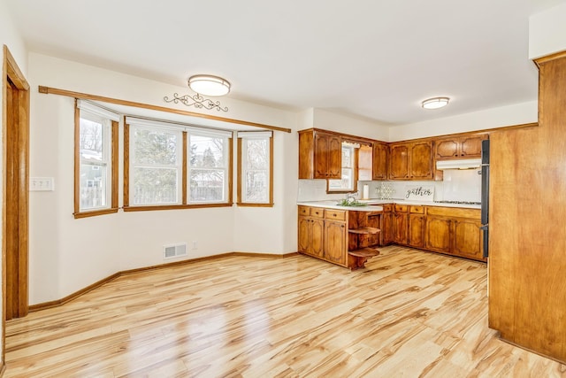 kitchen featuring visible vents, brown cabinetry, light wood-style flooring, light countertops, and under cabinet range hood