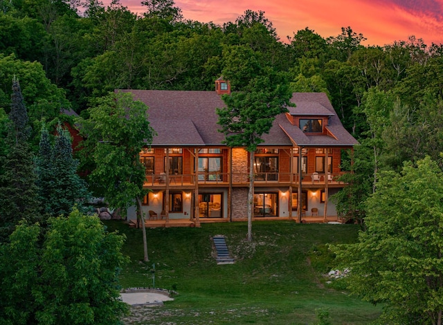 back of house at dusk with a patio area, stone siding, a view of trees, and a lawn