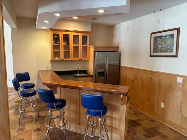 bar featuring a wainscoted wall, stone finish flooring, stainless steel refrigerator with ice dispenser, and a sink