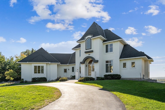 french country home with a shingled roof, curved driveway, a front yard, and stucco siding