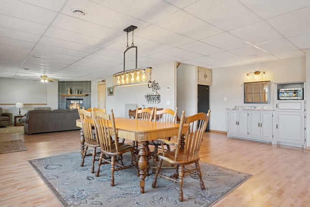 dining space with baseboards, light wood finished floors, a fireplace, a drop ceiling, and ceiling fan