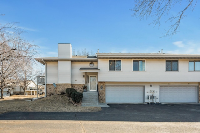 bi-level home featuring aphalt driveway, brick siding, a chimney, and an attached garage