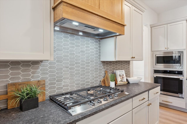 kitchen with custom exhaust hood, dark countertops, decorative backsplash, white cabinets, and appliances with stainless steel finishes