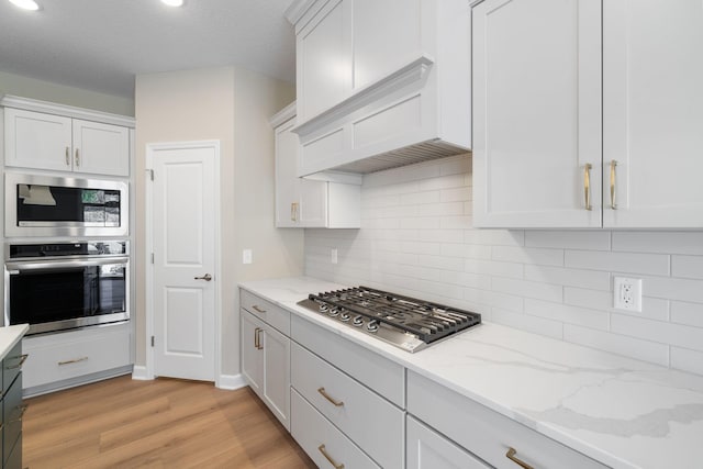 kitchen featuring light stone counters, stainless steel appliances, premium range hood, light wood-style floors, and tasteful backsplash