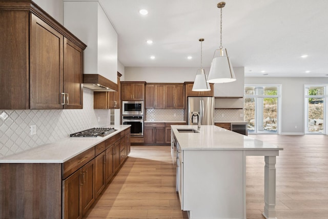 kitchen with a sink, light wood-style floors, wall chimney range hood, appliances with stainless steel finishes, and an island with sink