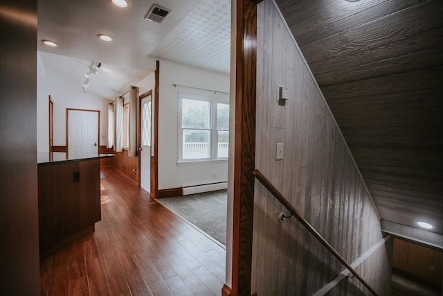 hallway featuring visible vents, dark wood-type flooring, wooden walls, a baseboard radiator, and vaulted ceiling