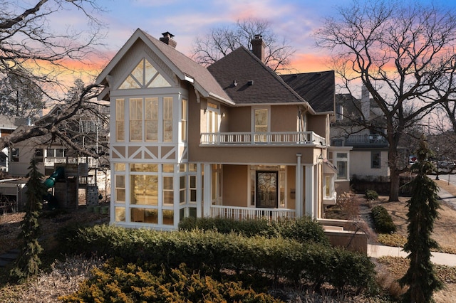 back of property at dusk with stucco siding, a porch, roof with shingles, a balcony, and a chimney