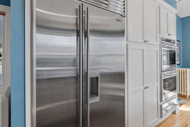 kitchen with light wood-type flooring, stainless steel appliances, white cabinets, and radiator heating unit