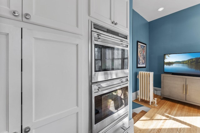 kitchen featuring radiator, baseboards, stainless steel double oven, white cabinetry, and light wood-type flooring