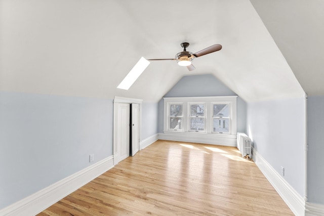 additional living space with light wood finished floors, baseboards, radiator heating unit, vaulted ceiling with skylight, and a ceiling fan