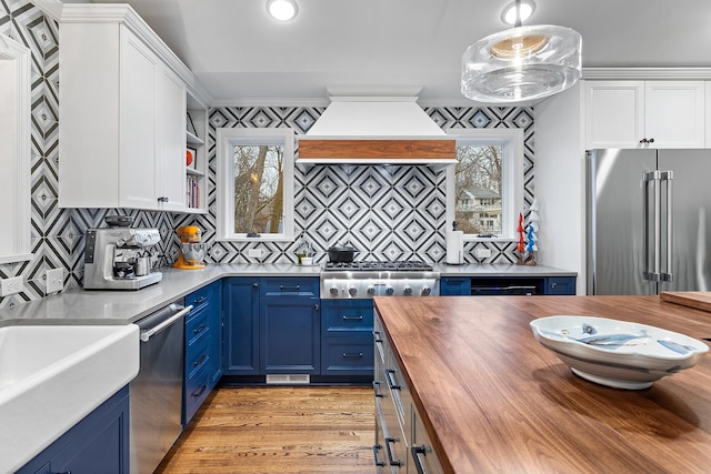 kitchen with stainless steel appliances, butcher block countertops, custom range hood, blue cabinetry, and crown molding