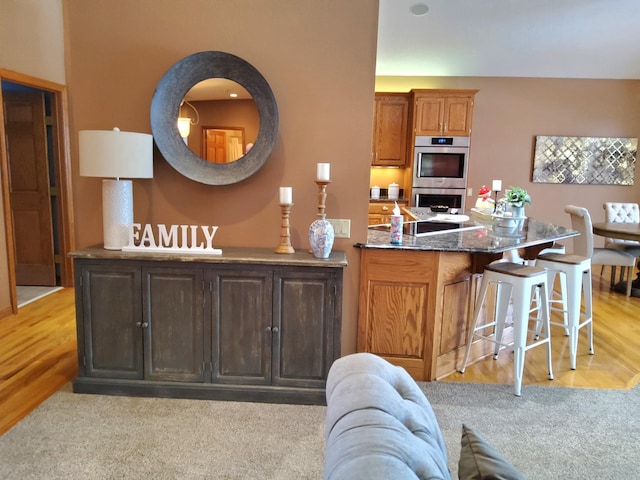 kitchen with stainless steel double oven, light carpet, a peninsula, dark stone counters, and a kitchen bar