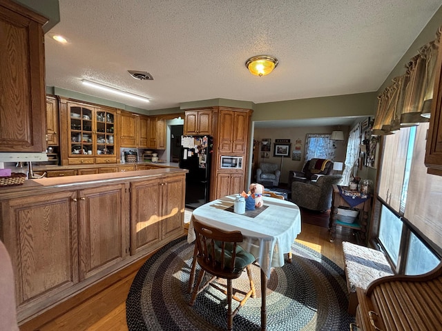 kitchen featuring stainless steel microwave, light wood-style flooring, brown cabinetry, freestanding refrigerator, and glass insert cabinets