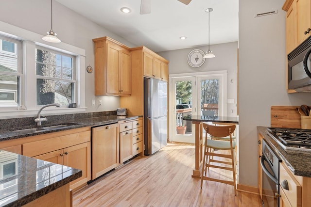 kitchen featuring dark stone counters, light brown cabinetry, a sink, light wood-style floors, and appliances with stainless steel finishes