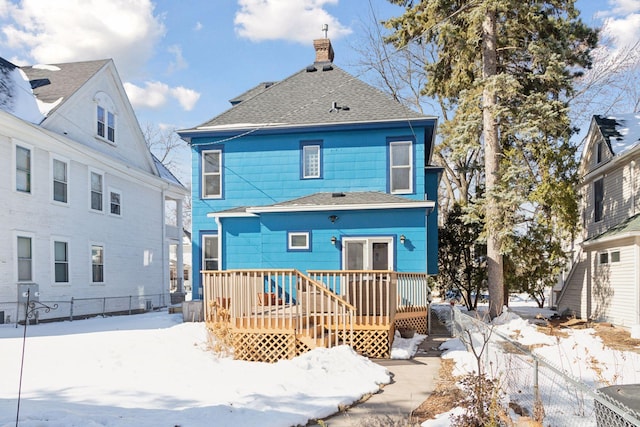 snow covered house featuring a chimney, a wooden deck, roof with shingles, and fence