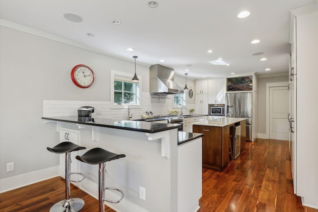 kitchen featuring tasteful backsplash, appliances with stainless steel finishes, wall chimney range hood, ornamental molding, and a kitchen bar
