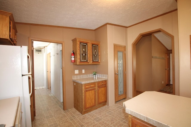 kitchen featuring brown cabinetry, ornamental molding, freestanding refrigerator, light countertops, and a textured ceiling