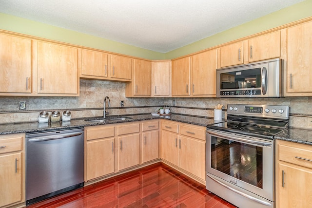 kitchen with dark stone countertops, light brown cabinets, a sink, decorative backsplash, and appliances with stainless steel finishes