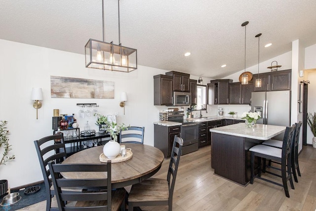 kitchen with dark brown cabinetry, lofted ceiling, hanging light fixtures, stainless steel appliances, and a sink