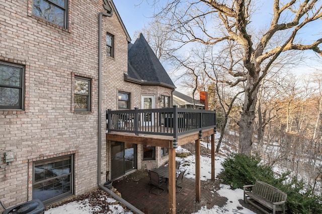 snow covered house with a patio, brick siding, roof with shingles, and a wooden deck