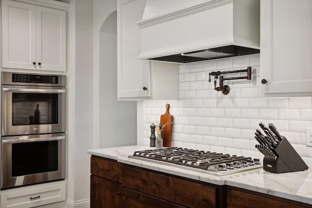 kitchen featuring light stone counters, stainless steel appliances, premium range hood, white cabinets, and tasteful backsplash