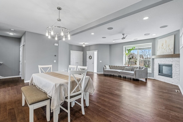 dining space with baseboards, dark wood-type flooring, a stone fireplace, and ceiling fan with notable chandelier