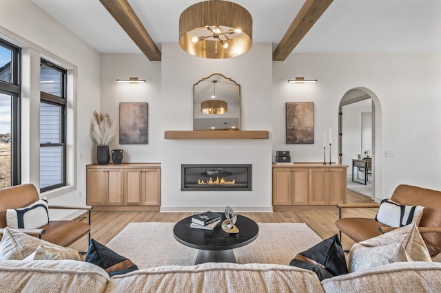 living room with arched walkways, a glass covered fireplace, beamed ceiling, and light wood-style flooring