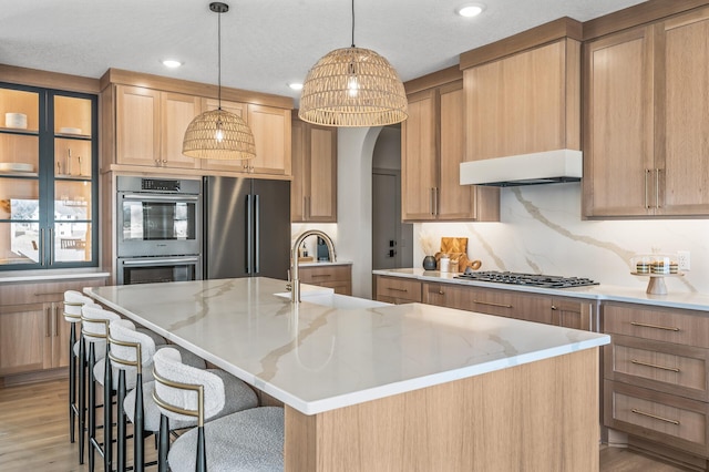 kitchen featuring a breakfast bar area, under cabinet range hood, stainless steel appliances, backsplash, and an island with sink