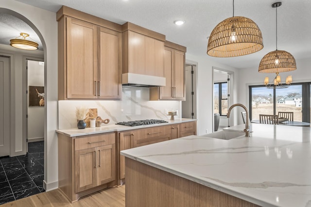 kitchen with under cabinet range hood, stainless steel gas cooktop, a sink, backsplash, and light stone countertops