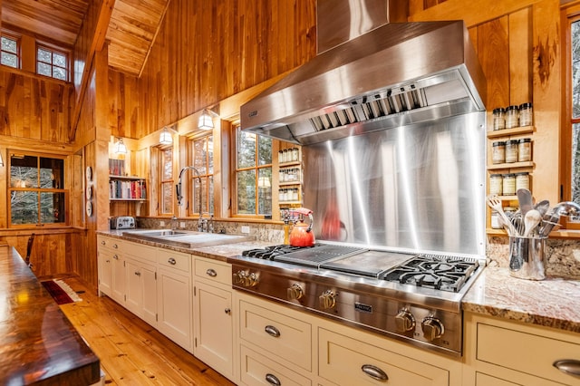kitchen with stainless steel gas stovetop, wood walls, wall chimney exhaust hood, a sink, and light wood-type flooring