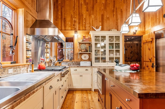 kitchen featuring wood walls, wall chimney exhaust hood, open shelves, light wood finished floors, and glass insert cabinets