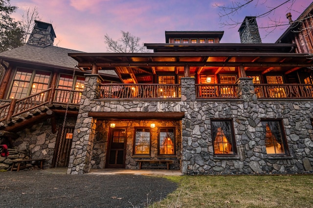 back of house at dusk with a chimney, a balcony, and stone siding