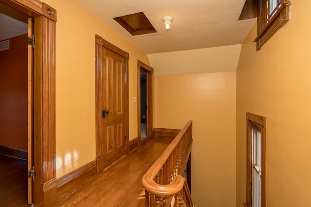 hallway featuring a textured ceiling, wood finished floors, an upstairs landing, baseboards, and vaulted ceiling