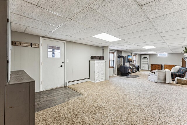 unfurnished living room with baseboard heating, a wood stove, carpet flooring, wood finished floors, and a drop ceiling