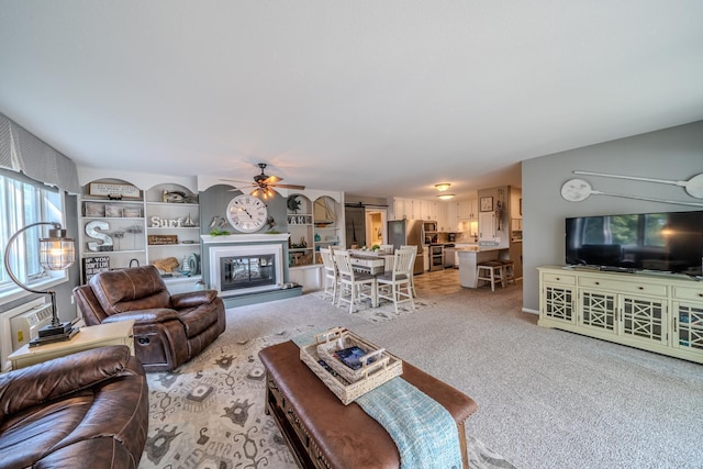 living room with a ceiling fan, a glass covered fireplace, light colored carpet, and built in features