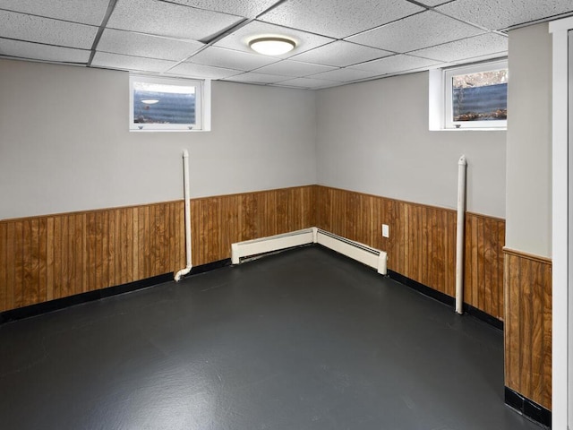 below grade area with a wainscoted wall, baseboard heating, a drop ceiling, and wooden walls