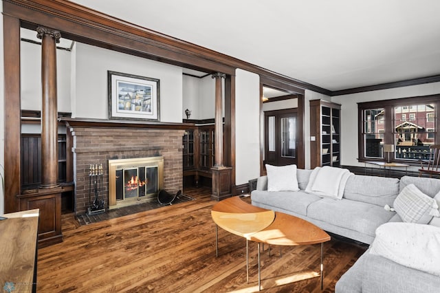 living area featuring wood finished floors, baseboards, a brick fireplace, decorative columns, and crown molding