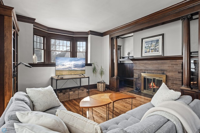 living area with wood finished floors, baseboards, a brick fireplace, decorative columns, and crown molding