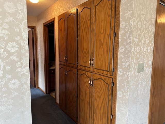 hallway with a textured ceiling, dark carpet, baseboards, and wallpapered walls