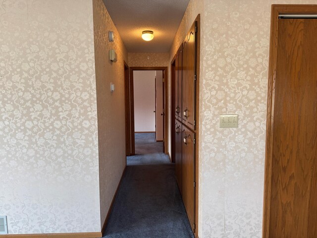 hallway with a textured ceiling, dark colored carpet, visible vents, and wallpapered walls