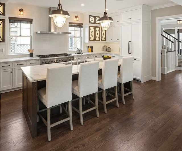 kitchen featuring wall chimney range hood, a kitchen island with sink, healthy amount of natural light, backsplash, and ornamental molding
