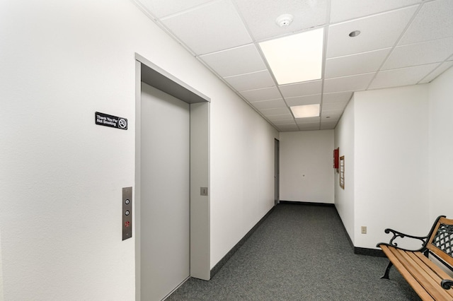 hall with dark colored carpet, a drop ceiling, elevator, and baseboards