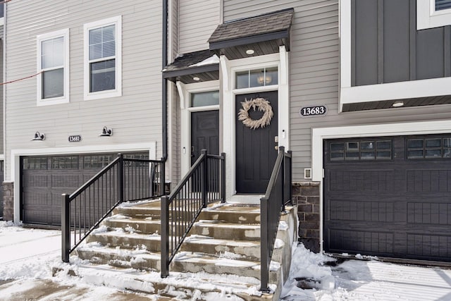 snow covered property entrance featuring an attached garage and a shingled roof