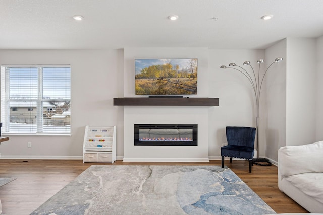 living area with a glass covered fireplace, baseboards, wood finished floors, and recessed lighting