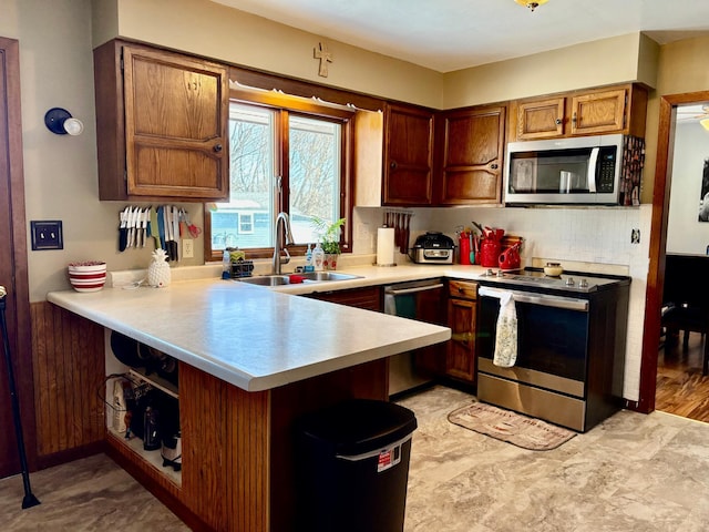 kitchen featuring a peninsula, a sink, light countertops, appliances with stainless steel finishes, and brown cabinetry