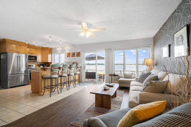 living room featuring a water view, light wood-style floors, ceiling fan, a textured ceiling, and wallpapered walls