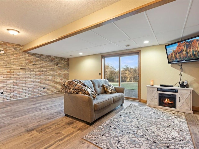 living room with a textured ceiling, brick wall, wood finished floors, a warm lit fireplace, and beamed ceiling