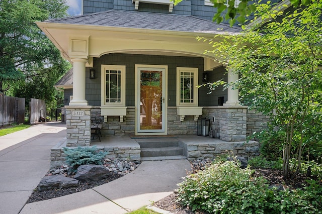 doorway to property featuring a porch, stone siding, roof with shingles, and fence