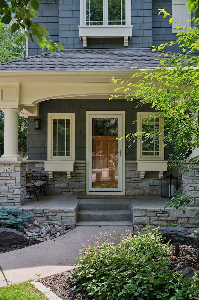 doorway to property with covered porch, stone siding, and roof with shingles